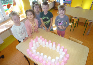 Pięcioro dzieci stoi przed stolikiem na którym ułożony jest uśmiech z białych i różowych kubeczków plastikowych.